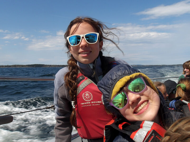The image shows two young women on a boat, both wearing sunglasses and life vests. They are smiling at the camera, and the background shows a body of water and a blue sky with some clouds. The overall impression is one of fun and adventure.